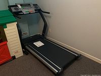 Image showing the full view of the treadmill from the running belt side, positioned in a corner against the walls indoors, manual resting on the treadmill belt.