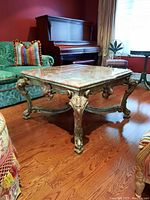 Side view of carved wood ornate leg and cross braces showing green patina and distressed finish, alongside hardwood flooring and surrounding furniture.