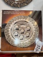 Photo of boxed antique silver plated round Passover plate with grapevine border and six labeled compartments.
