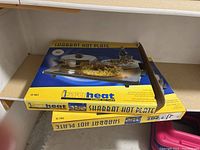 Two Israheat Shabbat hot plates in original boxes on a shelf. Yellow packaging with blue accents, labeled IS-901 and IS-702.