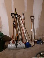 Photo showing standing shovels, brooms, and garden hand tool against unfinished drywall wall and concrete floor.