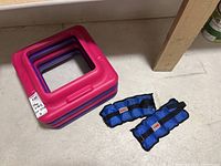 Four plastic step risers stacked, pink and purple, and two blue York ankle/wrist weights on floor near wall.