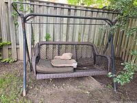 Front view of weathered outdoor swing bench with metal frame and wicker seat, including several cushions on the seat.