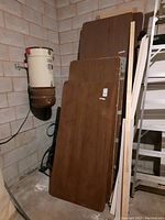 Stack of five wooden utility table tops leaned against a wall in a garage. Tables have brown wood grain surfaces with scratches and visible wear. Larger and smaller tabletops are stacked together.