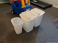 Three white plastic waste baskets. Two have attached lids, one without lid shows holes and scuffs near the bottom.