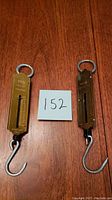 Two vintage brass fish scales placed side by side on a wooden surface with a note marked '152'