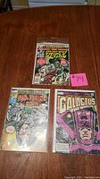 Three comic books on a wooden table with a pink numbered tag #74