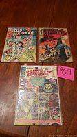 Three Silver Age comic books including Fantasy Masterpieces #1, Sub-Mariner #38, Nick Fury Agent of Shield #3, all on a wooden table with lot number 69 noted on a pink sticker.