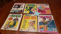 Six Cracked magazines arranged in two rows on a wooden table showing full front covers, with various cartoon illustrations and titles including 'Shut-Ups' edition and others.