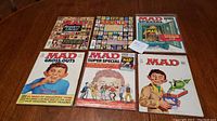 Top-down photo showing six vintage MAD magazines arranged in two rows on a table with various covers featuring caricatures and graphic cartoons.