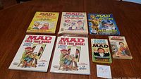 Lot of 7 Mad magazines and 2 Mad books laid out on wood surface, visible colorful illustrated covers with various humorous themes.