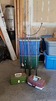 View of four fishing rods leaning against stacked plastic storage bins with two closed tackle boxes in front on the floor.
