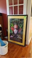 Framed Alice in Wonderland movie poster leaning against a wall near a window showing full view of the item and its size comparison to surroundings.