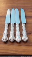 Four Birks sterling silver dinner knives with ornate Chantilly Lace patterned handles, laid out on wooden surface