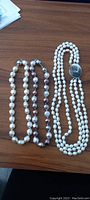 Top view of three pearl necklaces laid out on a wooden surface showing two white necklaces and one brown-toned pearl necklace.