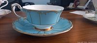 Side view of vintage turquoise blue Paragon teacup on matching saucer showing gold vertical stripes, gold scroll rim detailing, and embossed scale pattern on panels. Teacup handle also decorated with gold scrollwork.