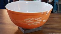 Front angle of vintage Pyrex mixing bowl showing orange exterior with beige wheat pattern and white interior on wooden surface.