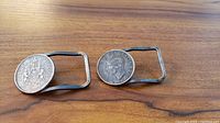 Two belt buckles on a wooden surface showing silver coins with historic designs mounted on rectangular metal frames.