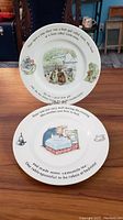 Two Wedgwood Beatrix Potter plates placed upright on a wooden surface showing the full illustrations and story text: one plate features Peter Rabbit in bed and the other Mrs. Tiggy-Winkle hanging laundry.