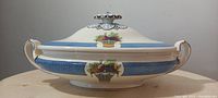 Side view of vintage Wedgwood Etruria Blue Directoire oval covered vegetable dish showing blue band and fruit/vegetable basket decorations on bowl and lid, with floral-shaped handle on lid.