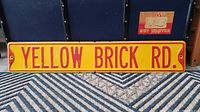 Front side of Yellow Brick Road street sign showing yellow background with red text lettering reading 'YELLOW BRICK RD.' mounted on a surface with visible mounting holes and minor surface marks.