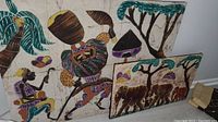 Three pieces of African cloth art shown together, two framed cloth panels and one tapestry, featuring people, animals, and nature from African themes