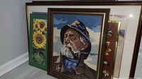 Lot of 7 framed art pieces leaning against a wall. Foreground piece is a needlepoint tapestry of an old man with a pipe.