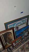 Six framed art pieces resting against a wall showing varied sizes and styles.