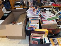 Photo showing large cardboard boxes and plastic bins filled with books, magazines, and VHS tapes stacked on floor with some trophies in background.