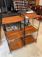 Pair of wooden and metal bookshelves with three shelves each, showing natural wood grain and black metal frames with decorative curves and caster wheels.