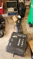 Full view of broadcast equipment lot on floor showing multiple tripods, heavy duty hard cases with labels, coiled cables, and cardboard boxes