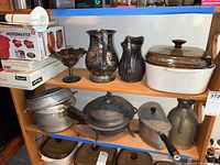 Wide view of shelf showing aluminum pots, decorative pitchers, a covered casserole dish, and a dark iridescent bowl.