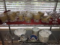 Shelves holding assorted dishware and glassware including wrapped white soup bowls, stacked white plates, ceramic bowls with floral and geometric patterns, and clear glass tumblers.