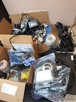 Photo showing a cardboard box filled with assorted electronics cables, power cords, adapters, and a calculator in plastic bag.