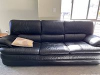 Front view of black leather couch with visible wear, showing three Cushioned backrest sections and armrests, with a beige pillow on the left side.