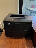 Photo of an HP LaserJet black and white printer on a wooden surface near a window, showing the front and top sides of the unit.