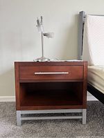 Frontal view showing drawer and open lower shelf of the night stand next to a bed frame, with a silver lamp on top.