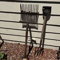 Three used garden tools leaning against an exterior wall: a large metal rake with long tines, a digging fork with wooden D-handle, and a hand cultivator with red handle.