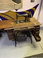 Full view of antique Singer sewing machine in wooden cabinet with multiple storage drawers and fold-out top sections, showing wear and surface patina.