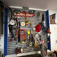 Photo of automotive tools and accessories hanging on a blue and gray metal wall rack with a Budweiser sign above and American flags attached.