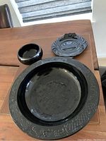 Four pieces of black Depression glass set on a wooden table, including bowls and plates with detailed embossed edges and patterns.