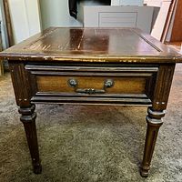 Side view of wooden end table showing single drawer with metal handle and tapered legs with carved details. Shows heavy surface wear and scratches.