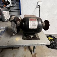 Buffalo Machine Tools bench grinder with wire brush and grinding wheel on each side, placed on a metal table with a power outlet in background.