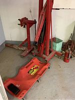 Photo showing red motor stand with adjustable mounting arm, red foldable engine hoist with hydraulic ram, and a large red plastic mechanic creeper with cushioned headrest on casters.