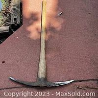 Full view of the pick tool showing the long wooden handle and metal iron head lying on a reddish surface.