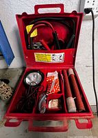Inside view of open red emergency kit showing all contents including two flares, jumper cables, large spotlight, gloves, pliers, and reflective cloth