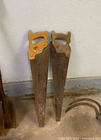 Photo of two vintage hand saws leaning against wall, showing wooden handles and rusted steel blades.