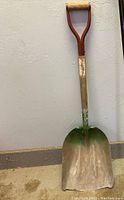 Photo of garden scoop shovel leaning against a wall showing wear on wooden handle and dirt on metal scoop.
