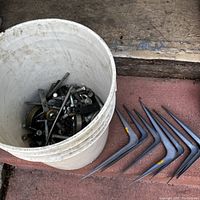 White bucket containing assorted screws, bolts, nuts, and hardware alongside grey metal L-shaped shelf brackets
