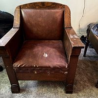 Front view of vintage wooden armchair with quarter sawn oak arms and heavily worn brown leather upholstery with visible damage on the seat cushion and some wear on the wood.